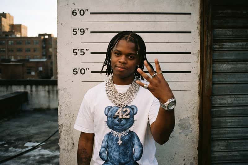 42 Dugg standing against a plain backdrop, wearing casual 42 Dugg standing against a plain backdrop, wearing casual streetwear, with height reference markers visible on the wall behin